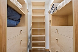 A narrow walk-in closet with wooden shelving and drawers, a central folding ladder, and a blue pillow on the left shelf.
