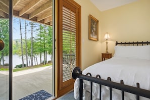 A bedroom with a bed, a nightstand, a lamp, a window with blinds, and a door leading to an outdoor area with trees and a view of a body of water.