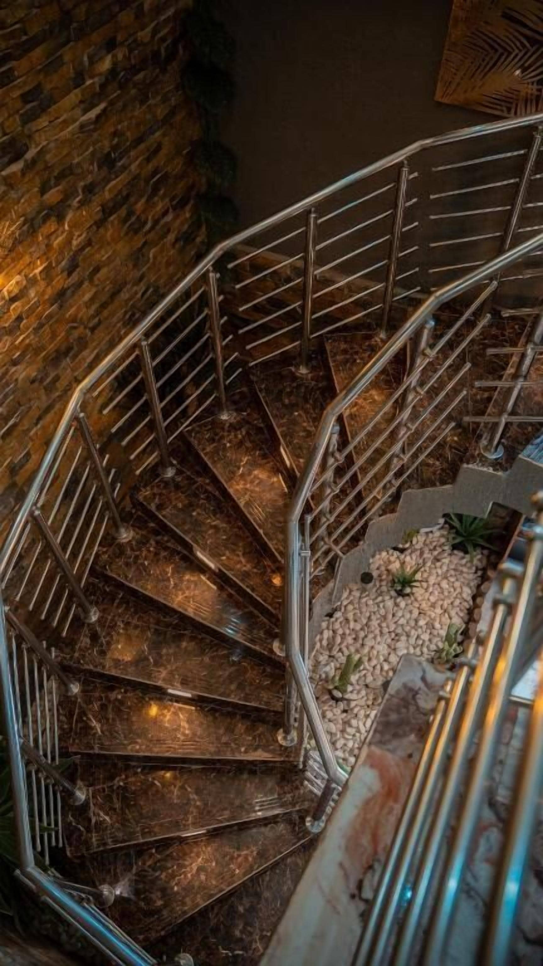 Eine Wendeltreppe mit Metallgeländern und Stufen aus dunklem Material, umgeben von einer Steinmauer und einer kleinen Fläche mit weißen Kieselsteinen und Pflanzen.