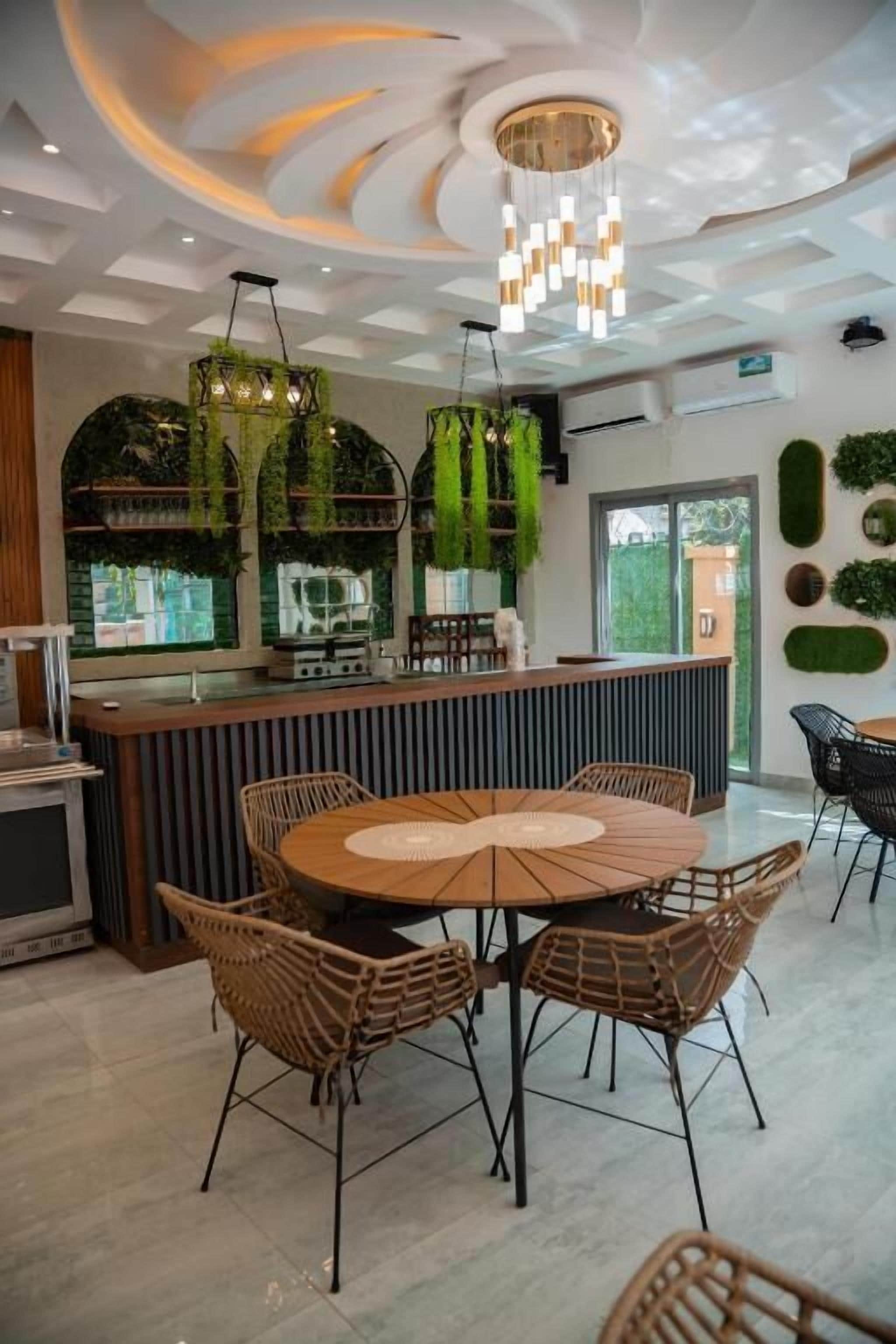 Ein modernes Interieur mit einem runden Esstisch, umringt von Korbstühlen, einer Bar mit Pflanzen und einer Decke mit einer besonderen Leuchte.