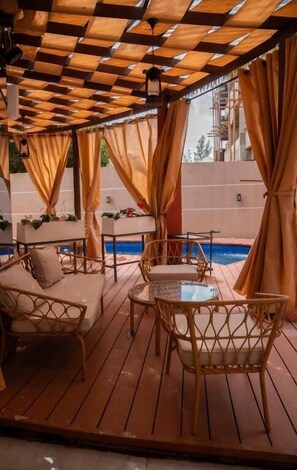 Un área de estar exterior con mobiliario de mimbre y una piscina al fondo.