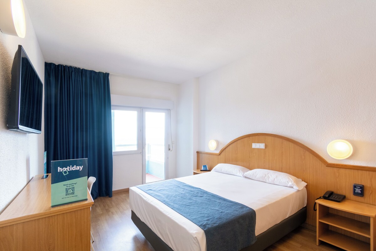 Habitación de hotel con cama, mesitas de noche, escritorio con un cartel, televisor y ventana con cortinas.