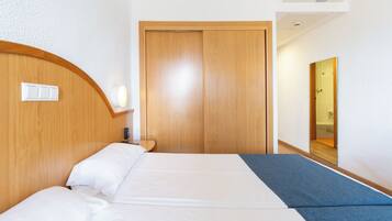 Een hotelkamer met een houten hoofdeinde, een bed met witte lakens, een nachtkastje, een wandlamp en een houten deur.