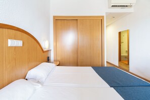 Una habitación de hotel con cabecera de madera, cama con ropa de cama blanca, mesita de noche, lámpara de pared y puerta de madera.