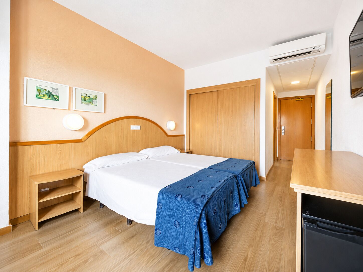 A hotel room with a bed, bedside tables, a desk, an air conditioning unit, and a door.