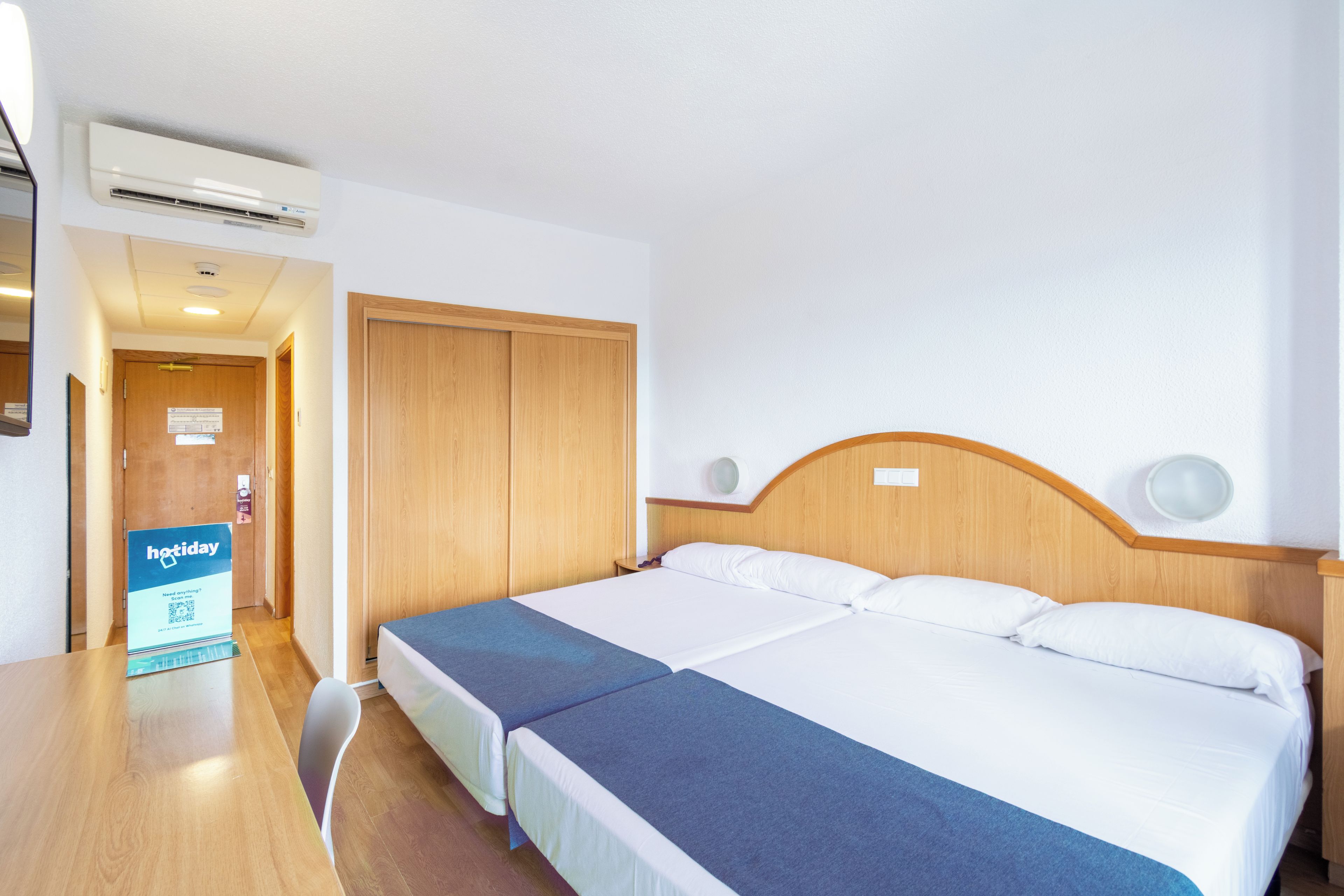 A hotel room with a double bed, a desk, and a chair. There is a sign on the desk and a door with a sign on it.