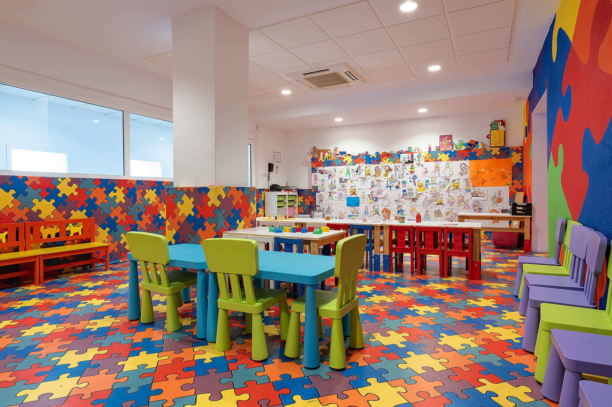 A children's playroom with colorful puzzle flooring, tables, chairs, and a wall decorated with children's artwork.