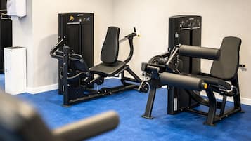 Uma academia com equipamentos de exercício, um dispensador de álcool em gel fixado na parede e uma máquina branca.