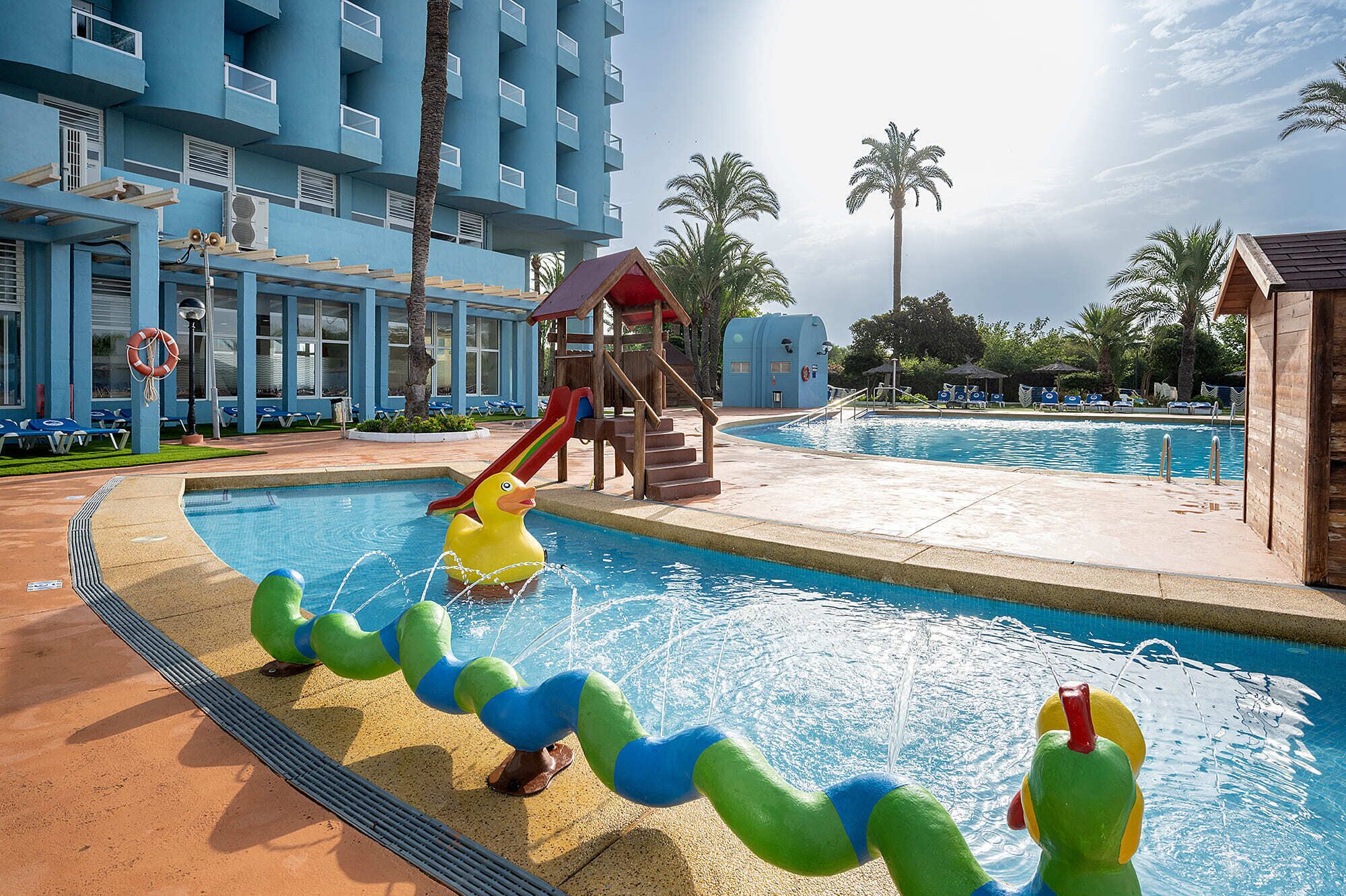 A swimming pool with a slide and a lifeguard tower, surrounded by palm trees and lounge chairs.