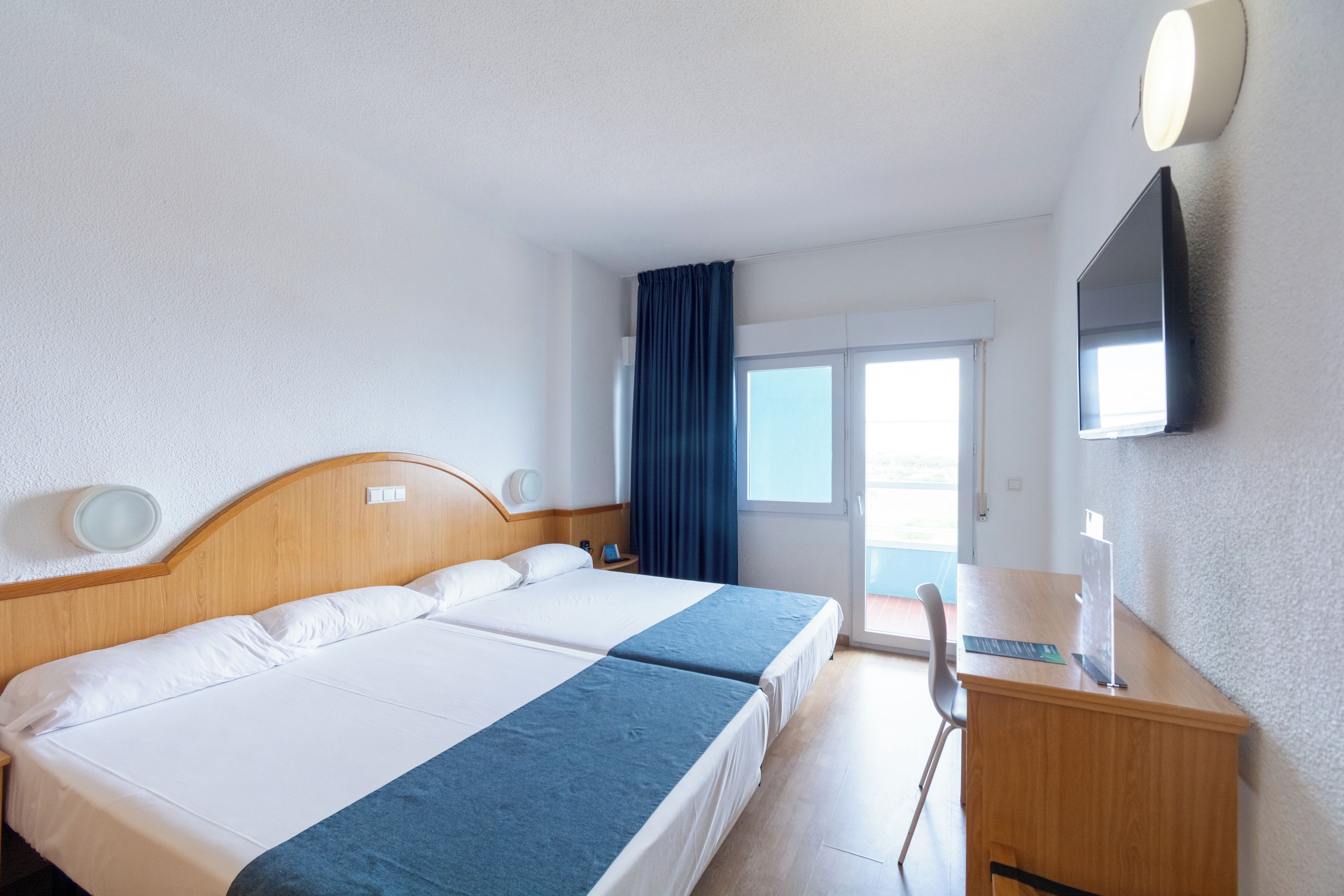 A hotel room with a double bed, a desk, a chair, a TV mounted on the wall, and a window with curtains.