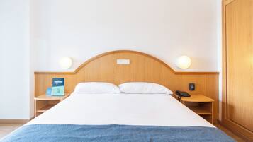 Habitación de hotel con cabecera de madera, dos lámparas de noche, cama con ropa blanca, una manta azul, mesita de noche con un teléfono y un letrero, y un armario de madera.