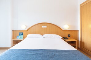 Habitación de hotel con cabecera de madera, dos lámparas de noche, cama con ropa blanca, una manta azul, mesita de noche con un teléfono y un letrero, y un armario de madera.