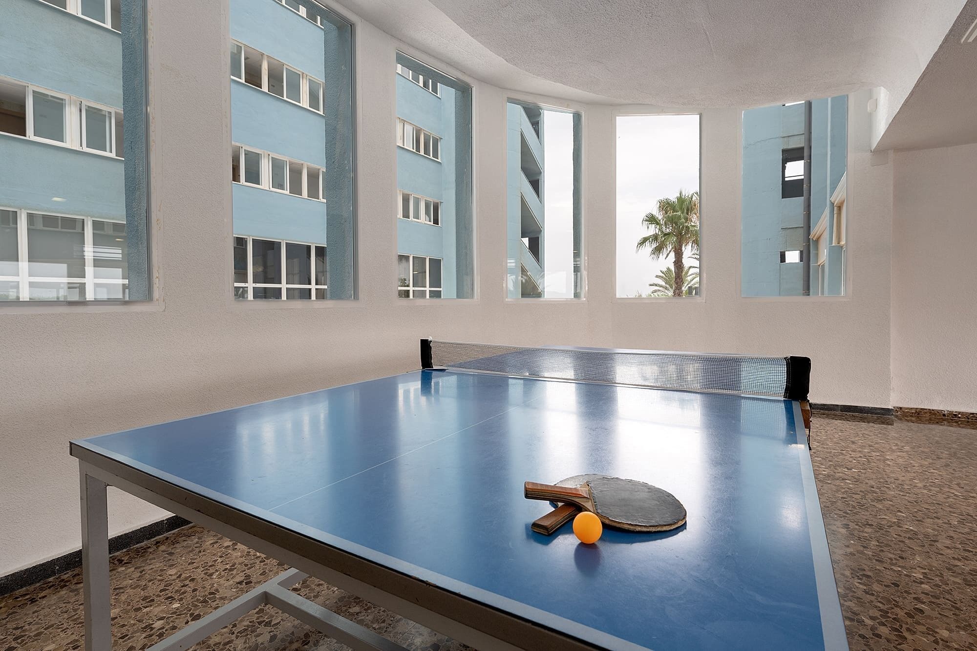 A ping pong table with a paddle and ball in a room with large windows and a view of a building.