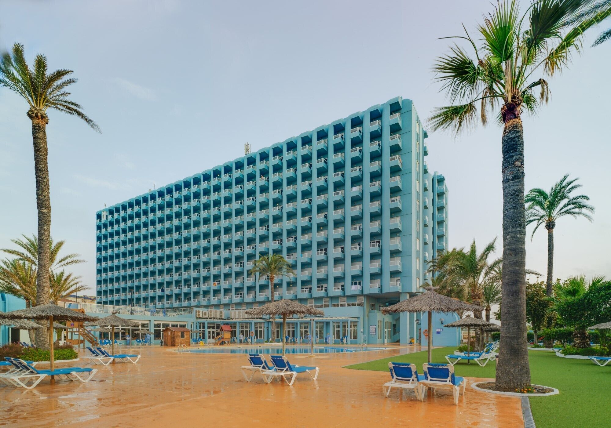 Een hotel met een groot blauw gebouw, een zwembad en palmbomen.
