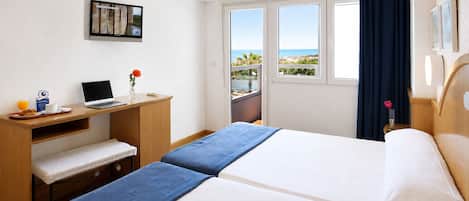 Habitación de hotel con cama, escritorio con computadora portátil, mesita con taza y jugo de naranja, y vista al mar por la ventana.