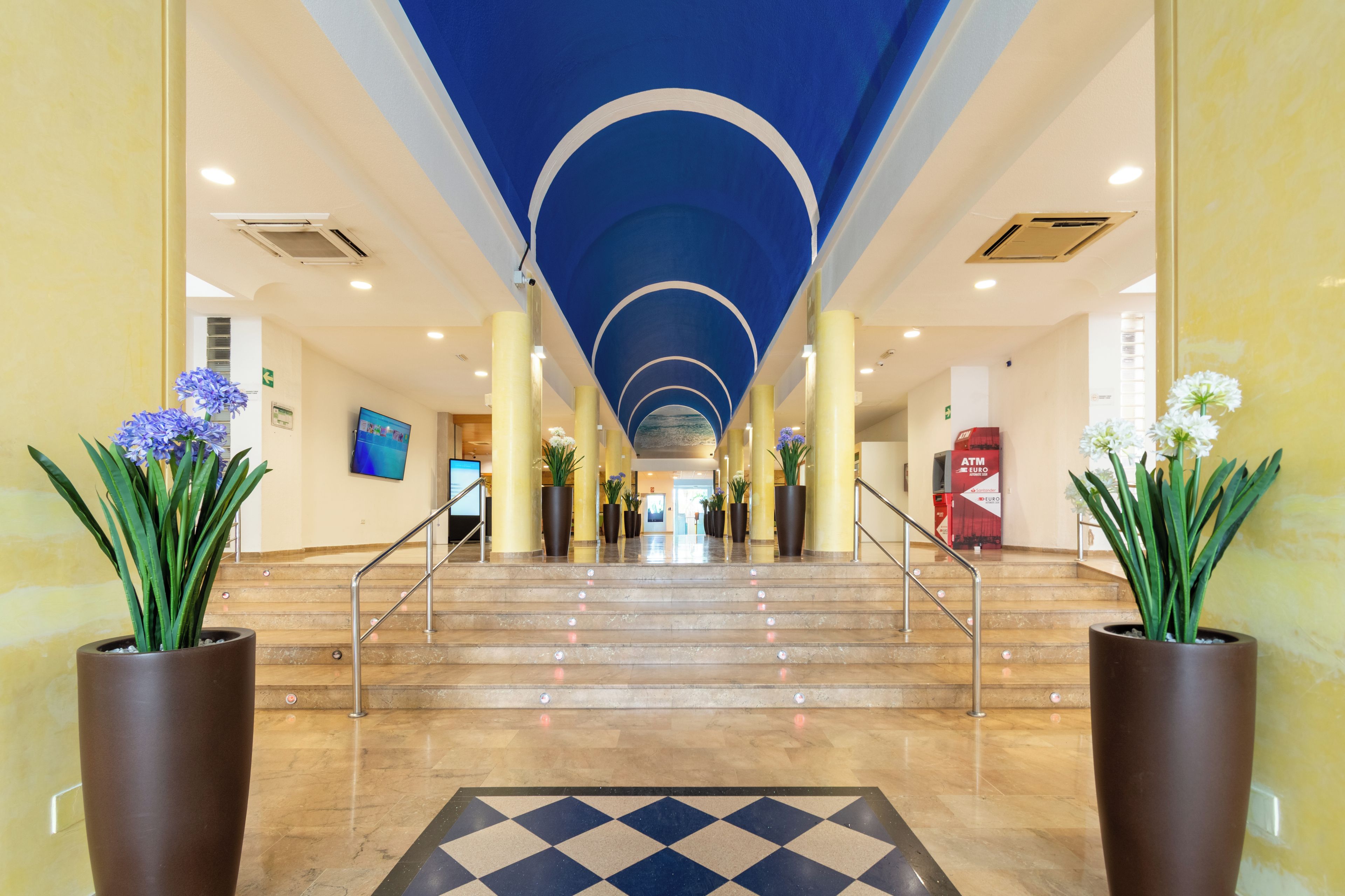 Um hall de hotel com uma escadaria de mármore, grandes vasos de plantas e um padrão de piso quadriculado.