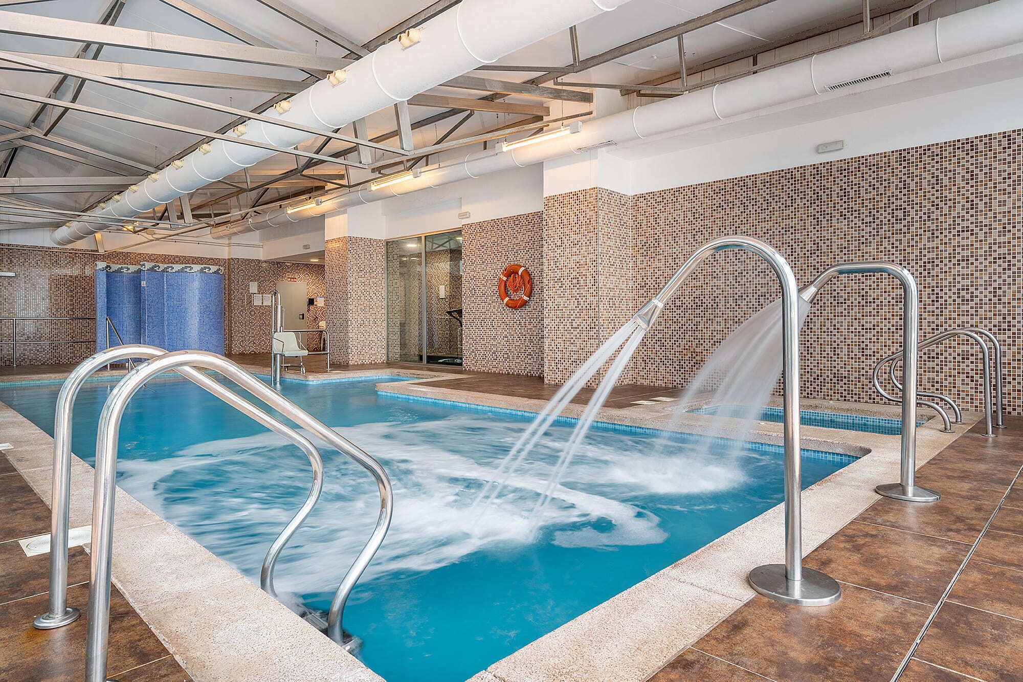 Una piscina interior con una fuente de agua cristalina, rodeada de paredes de azulejos y barandillas de metal.