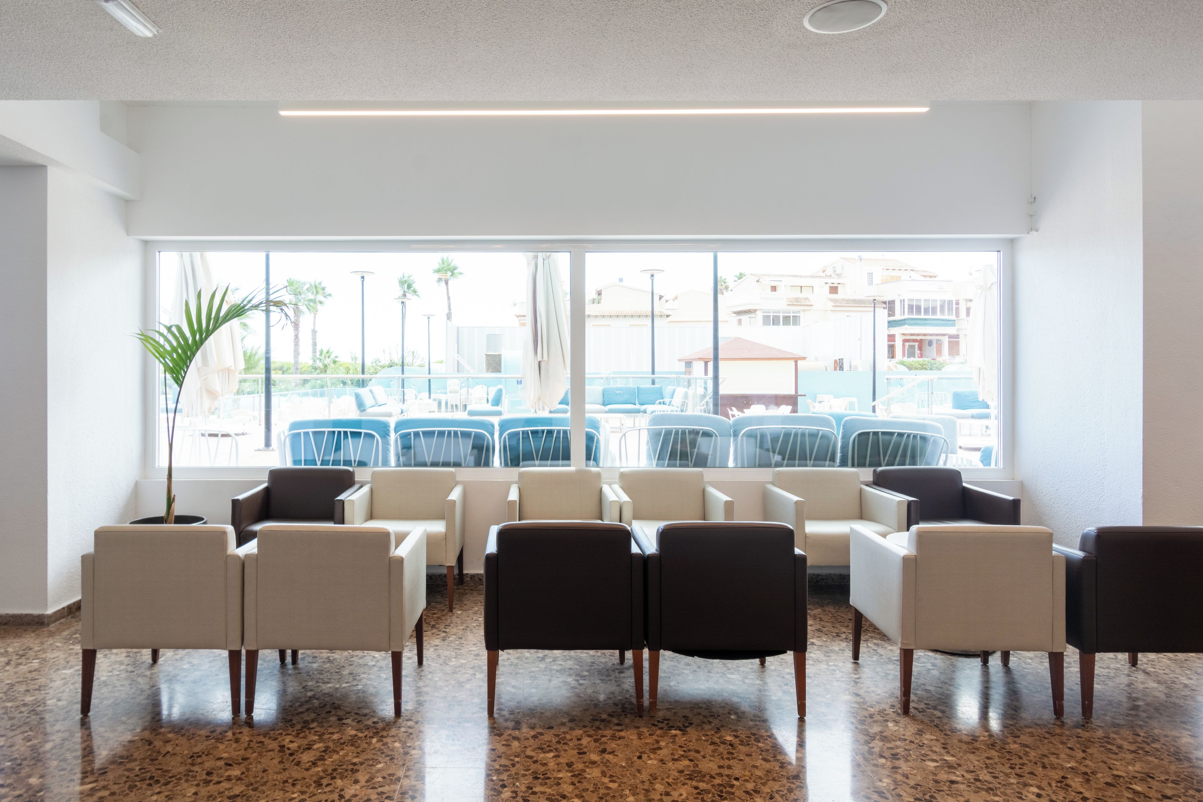 A waiting area with chairs arranged in rows, large windows with a view of outdoor seating, and a ceiling with recessed lighting.