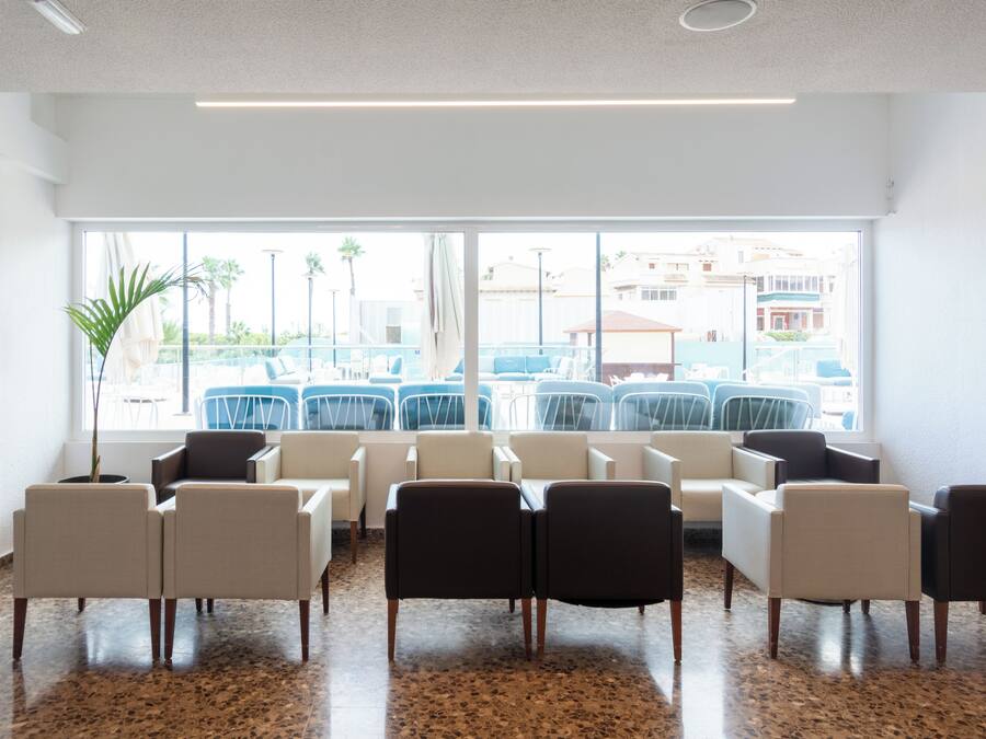 A waiting area with chairs arranged in rows, large windows with a view of outdoor seating, and a ceiling with recessed lighting.