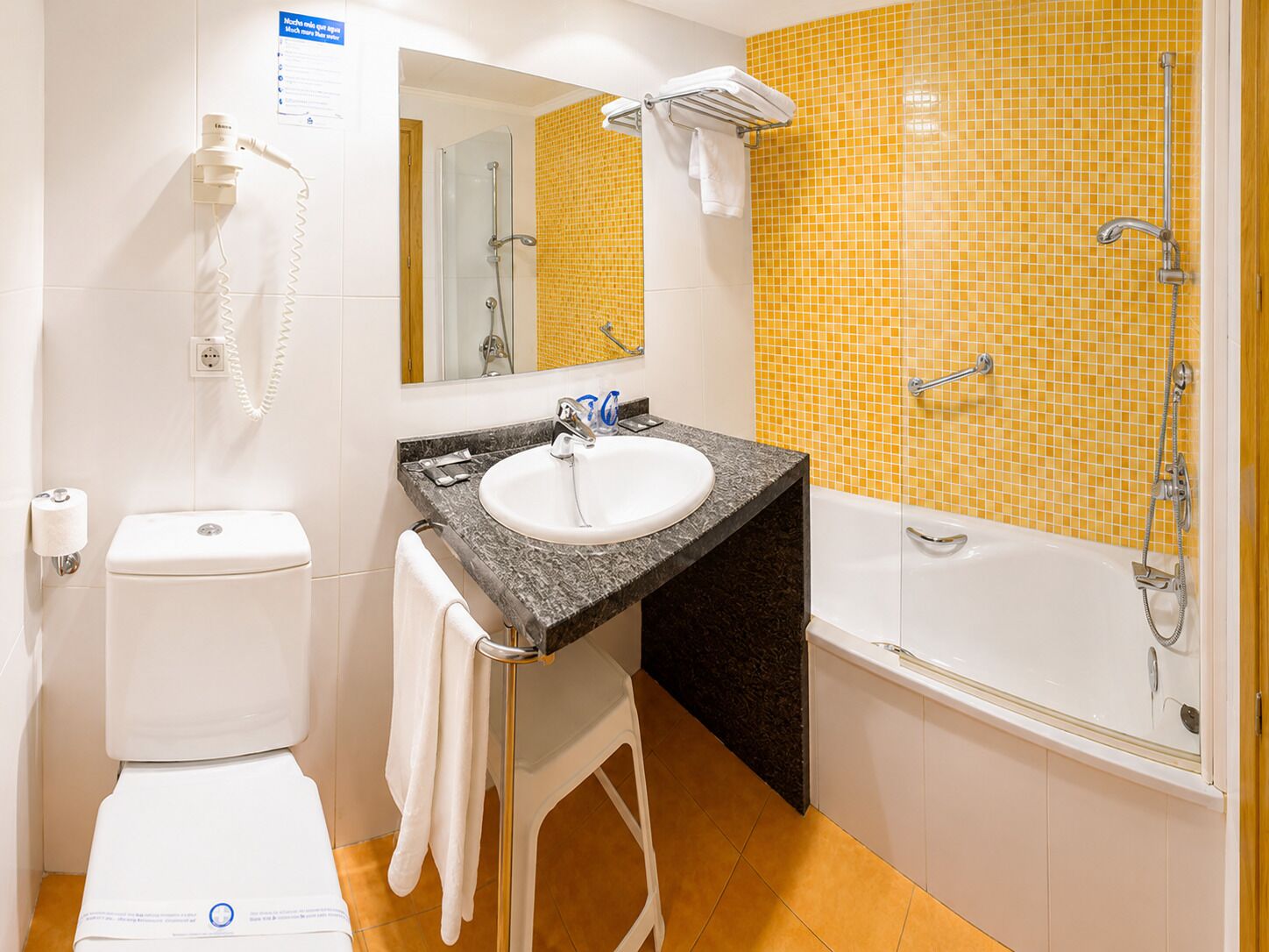 Een badkamer met een wit toilet, een wastafel met een marmeren blad, een douche met een gele betegelde wand en een handdoekhouder.