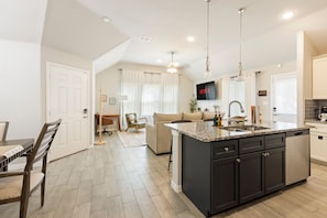 A modern kitchen with a central island, a dining area with a wooden table and chairs, and a living space with a sofa and a TV.