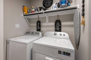 A laundry area with two white front-loading washers and dryers, a shelf above with cleaning supplies, and decorative wall hangings.