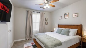 A hotel room with a bed, a ceiling fan, a TV mounted on the wall, and a window with curtains. - Cozy Home | 3BR Near Downtown FW | Sleeps 8 (Fort Worth)