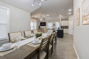 A modern kitchen with a central island, dining area with a long table, and a living room with a TV.