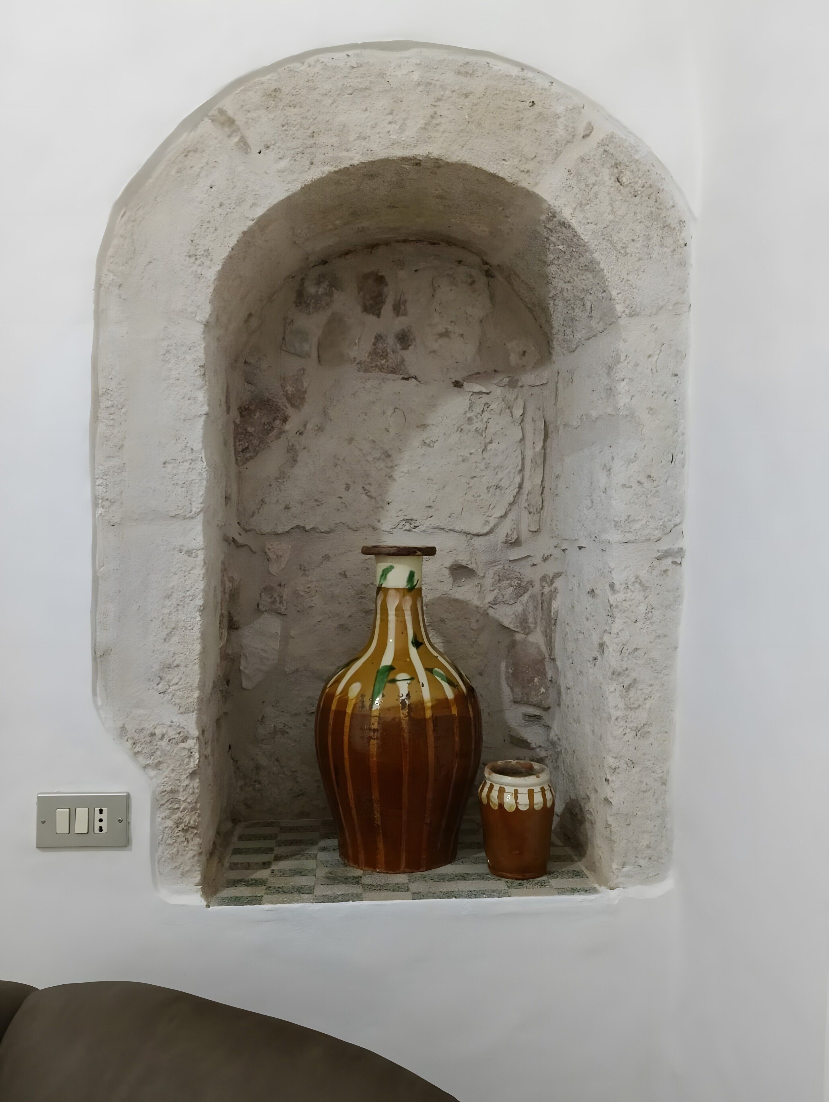 Eine weiße Wand mit einem steinernen Bogen. Im Bogen steht eine große Vase mit vertikalen Streifen und ein kleinerer Topf.