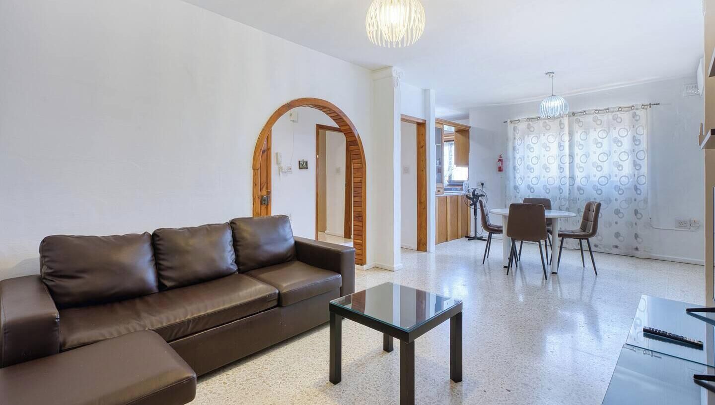 A living room with a brown leather sofa, a glass coffee table, and a dining area with a table and chairs.