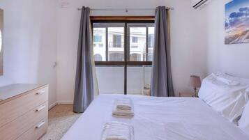 A hotel room with a bed, a dresser, a window with curtains, and a painting on the wall.