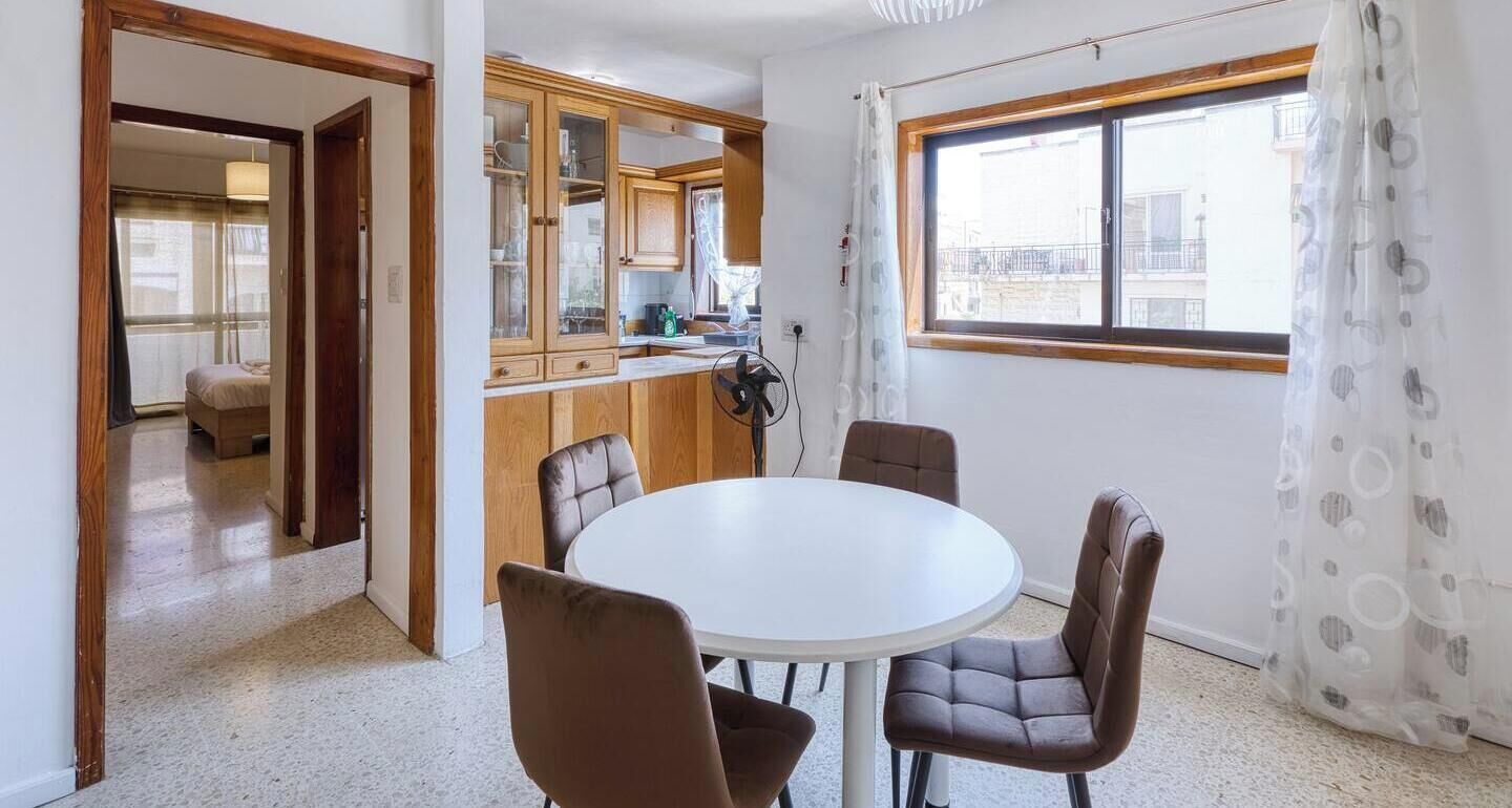 A compact kitchen with wooden cabinets, a dining table with four chairs, and a window with a view of buildings.