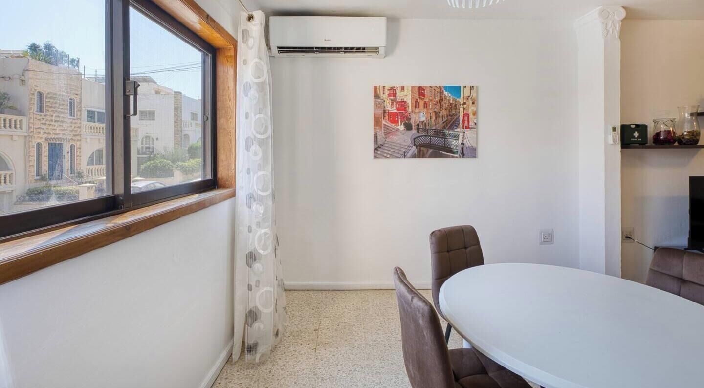 A room with a window, an air conditioning unit, a table with chairs, a picture on the wall, and a small shelf with items.