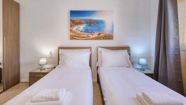 A hotel room with two beds, a painting of a coastal landscape, and a curtain on the window. - Beautiful 2BR Apartment in Swieqi with Balcony (Swieqi)