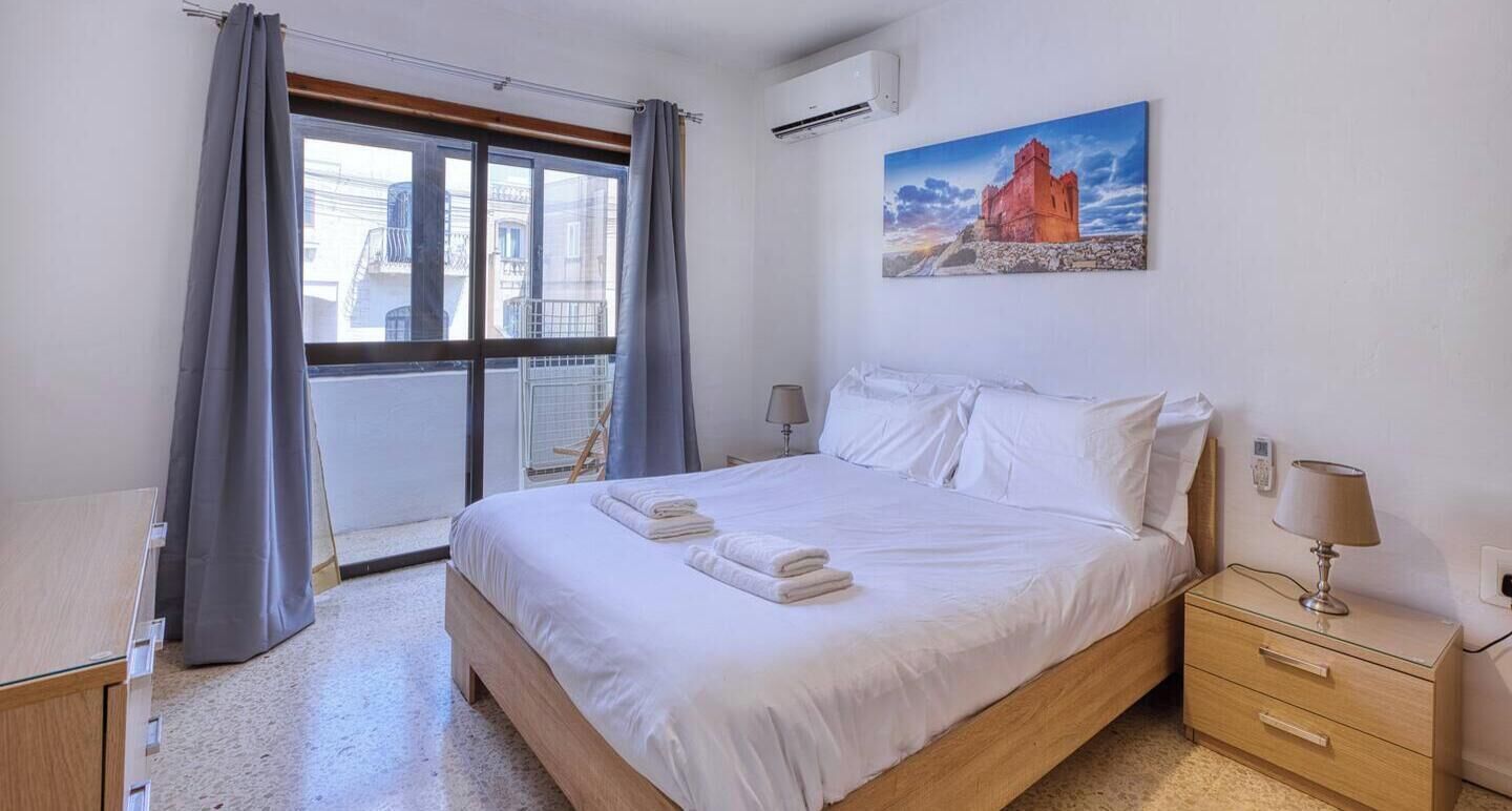Een slaapkamer met een bed, nachtkastje, lamp, schilderij en een raam met gordijnen.