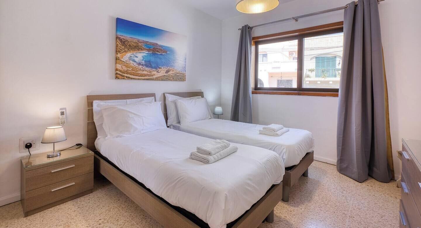 Une chambre d’hôtel avec deux lits, une table de chevet avec une lampe, un tableau représentant un paysage côtier et une fenêtre avec des rideaux.