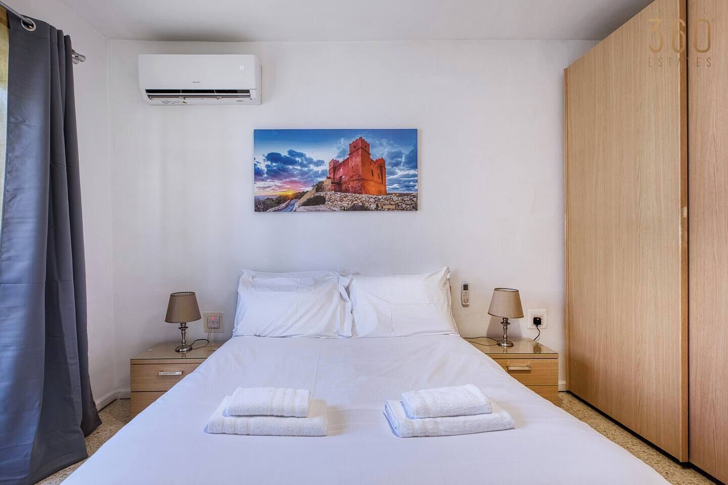 A hotel room with a bed, two bedside tables with lamps, a wall-mounted air conditioner, a painting of a castle, and a wardrobe.