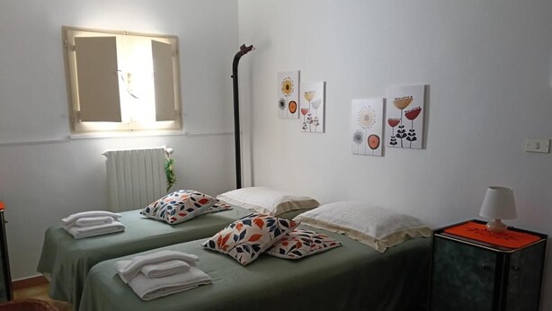 A room with two beds, each with a pillow and a folded towel. There is a radiator, a wall-mounted light, and a small table with a lamp.