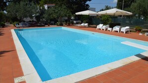 A rectangular swimming pool with a tiled edge, surrounded by a tiled deck. There are white lounge chairs and umbrellas, and the area is landscaped with trees and shrubs.