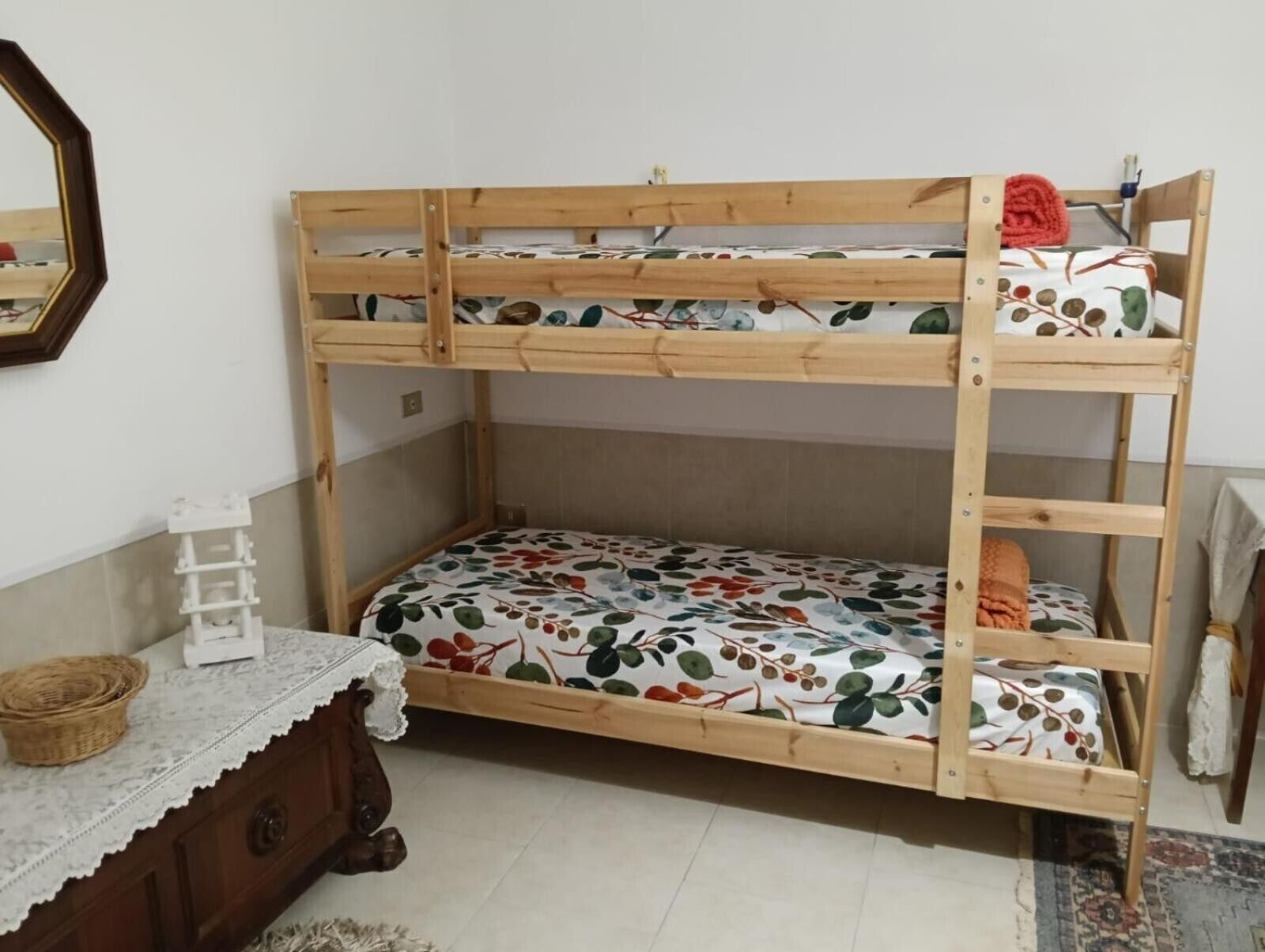 Ein Stockbett mit floraler Bettdecke, ein kleiner weißer Nachttisch mit einem Korb und eine Holzkommode.