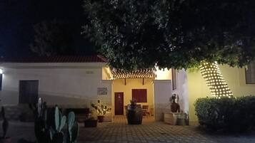 Un patio con un camino pavimentado, plantas en macetas, un banco y un edificio con una puerta roja y un cartel arriba.
