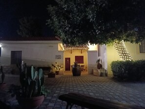 A courtyard with a paved walkway, potted plants, a bench, and a building with a red door and a sign above it.