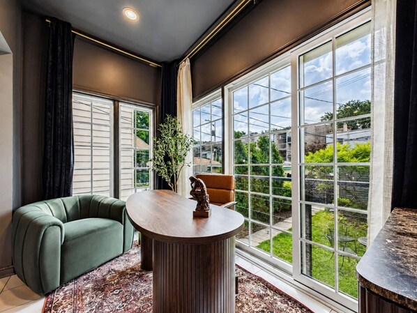 A modern interior with a green sofa, a round wooden table, and large windows offering a view of greenery and buildings.