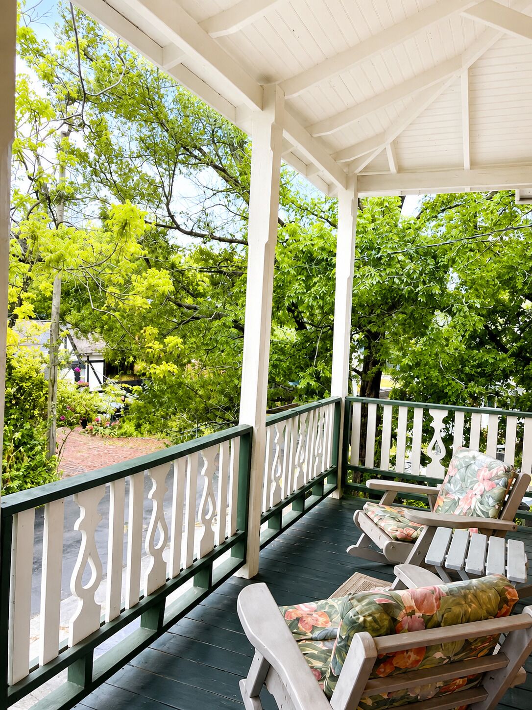 A covered porch with two wooden chairs, a railing, and a view of trees and a brick pathway.