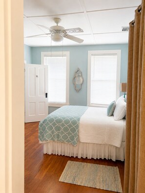 Une chambre à coucher comprenant un lit, un ventilateur de plafond, deux fenêtres avec stores, une porte et un tapis.