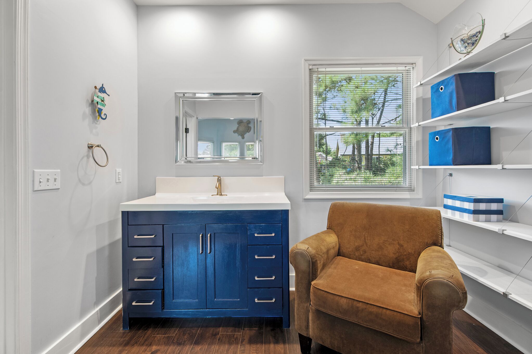 A bathroom with a white vanity, a blue cabinet, a brown chair, and a window with blinds.