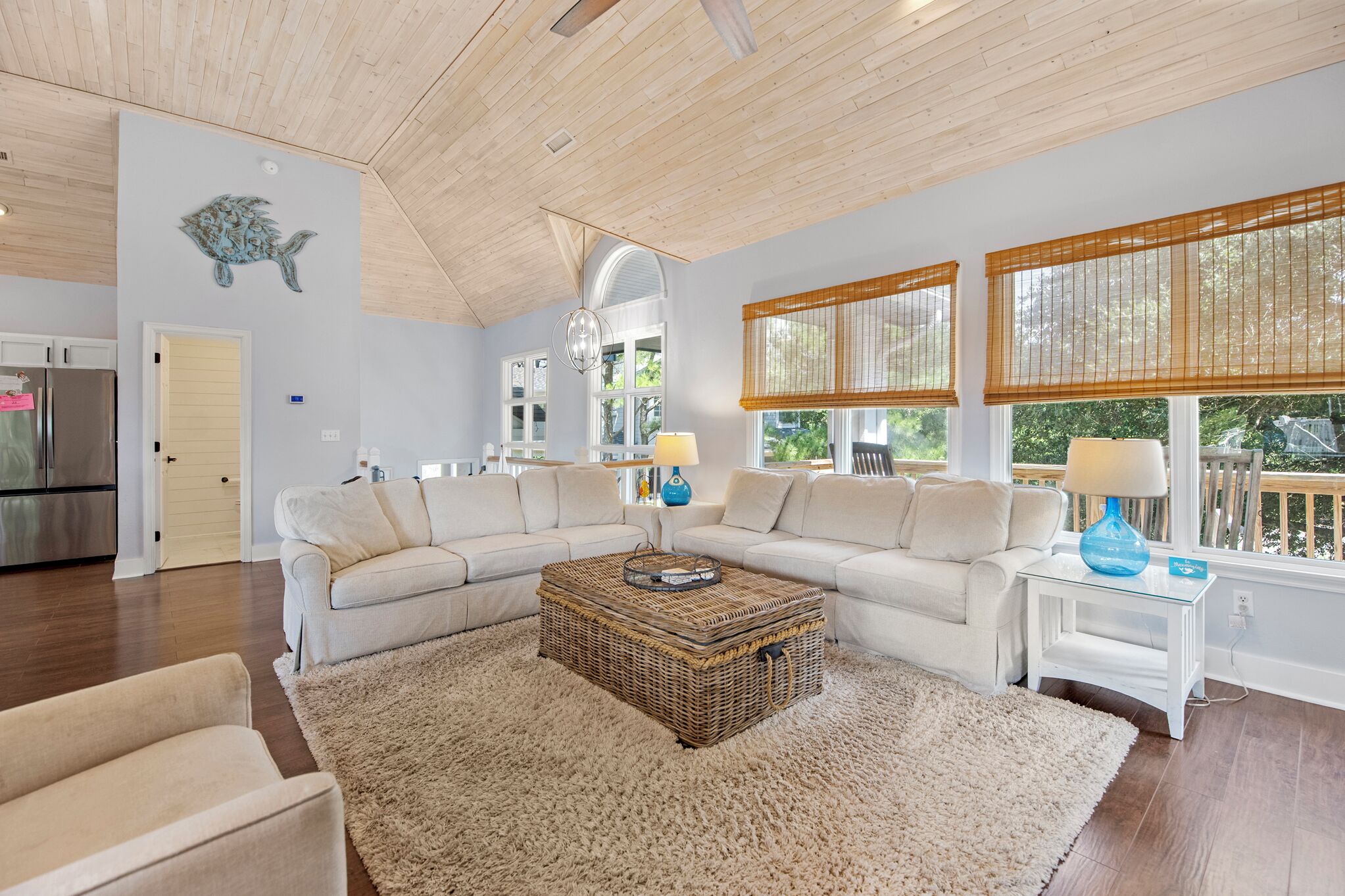 A spacious living room with a large L-shaped sectional sofa, a woven coffee table, and a fluffy rug. There are two side tables with lamps, a wooden ceiling, and large windows with blinds.