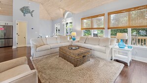 Ein geräumiges Wohnzimmer mit einer großen, L-förmigen Couch, einem gewebten Couchtisch und einem flauschigen Teppich. Zwei Nachttische mit Lampen, eine Holzboden und große Fenster mit Jalousien sind ebenfalls vorhanden.