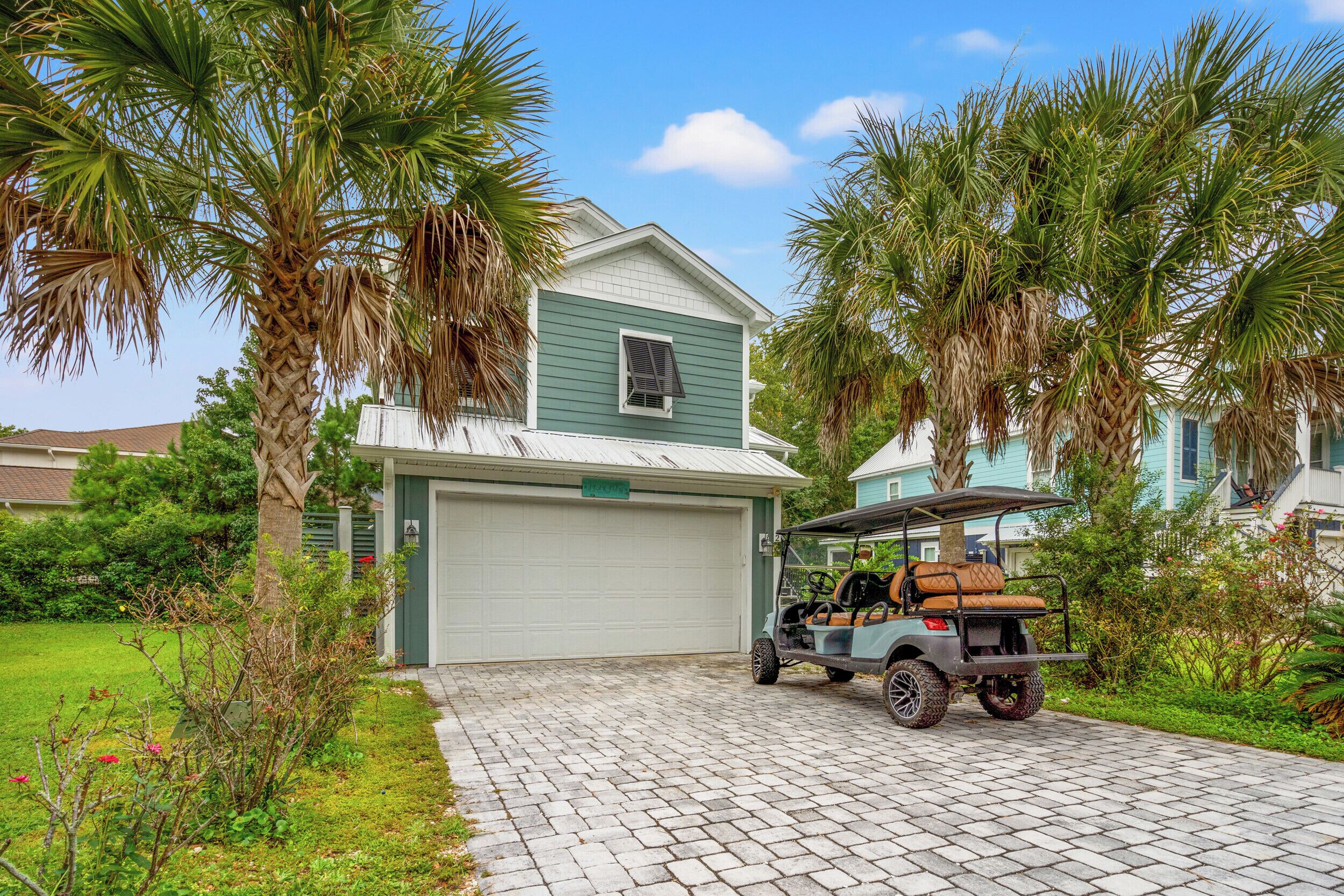 A two-story house with a garage, surrounded by palm trees and a paved driveway.