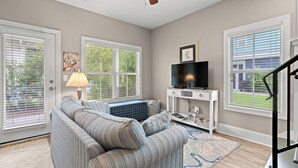 A living room with a sofa, a television on a stand, a lamp, and a large window with blinds. - Green Sandlapper: Golf cart included, North Litchfield Pawleys Island (Pawleys Island)